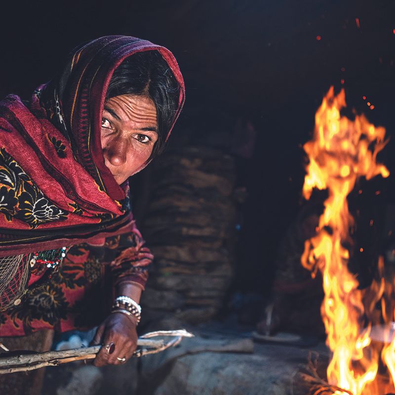 Eine Frau in traditioneller pakistanischer Kleidung mit Kopftuch beugt sich über ein offenes Feuer in dunkler Umgebung